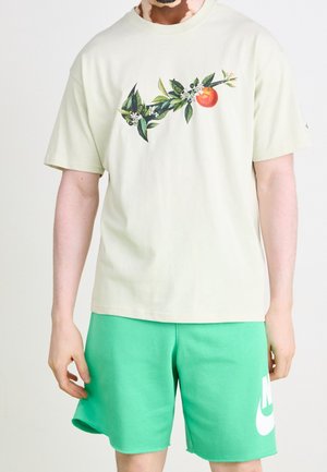 Ljusgrön bomulls-t-shirt med botanisk grafisk design med orange och blad. I kombination med gröna shorts som visar en vit logotyp.