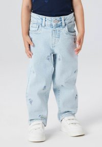 Bērns ar gaišziliem džinsiem ar maziem violetiem ziedu rakstiem un baltām velcro kedām, stāv uz vienkāršas fona.