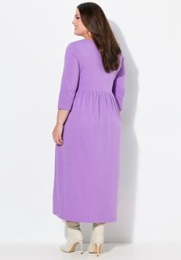 Gaun maxi lengan panjang berwarna ungu dengan bodice yang pas dan pinggang yang berkumpul. Kain halus, leher bulat, dipadukan dengan sepatu bot hak tinggi berwarna beige.