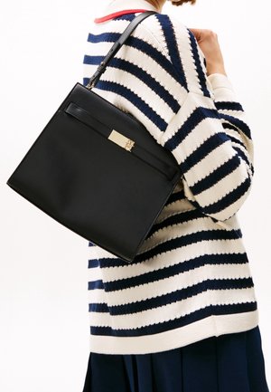 Personne portant un sac à bandoulière en cuir noir avec un fermoir doré, vêtue d'un pull crème et bleu rayé et d'une jupe sombre.