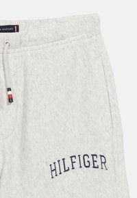 Ανοιχτό γκρι φόρμα παντελόνι με τη λέξη "HILFIGER" κεντημένη σε σκούρο μπλε κοντά στην τσέπη και κορδόνι με κόκκινες, λευκές και μαύρες λεπτομέρειες.