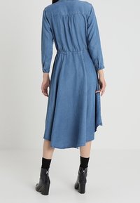Robe chemise en jean à manches longues, taille cintrée et jupe évasée. Présente un lavage bleu clair et un ourlet brut. Bottines noires à texture brillante.