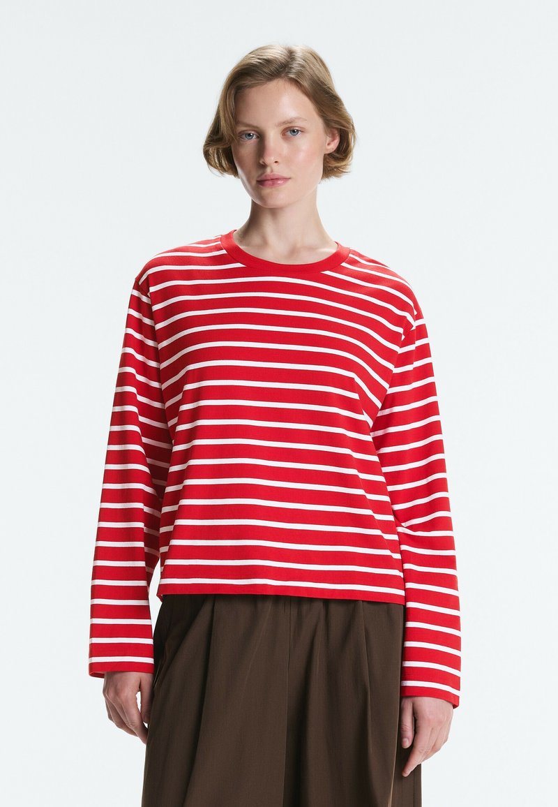 Jeune femme aux cheveux courts portant un t-shirt à manches longues rayé rouge et blanc et un pantalon marron foncé, debout devant un fond uni.