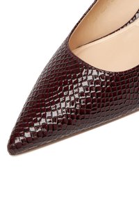 Czółenka na wysokim obcasie z ostro zakończonym noskiem w burgundowej lakierowanej skórze z teksturowanym wzorem przypominającym skórę węża. Odkryte boki i gładkie wnętrze.