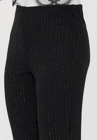Pantalons noirs ajustés avec de fines rayures verticales argentées portés avec un haut blanc doté d'une finition en dentelle noire.