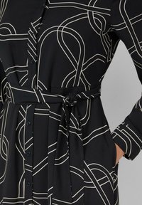 Zwarte jurk met wit abstract lijnpatroon, met een knoopsluiting aan de voorkant, lange mouwen en een gestrikte tailleband. Soepele stoftextuur.