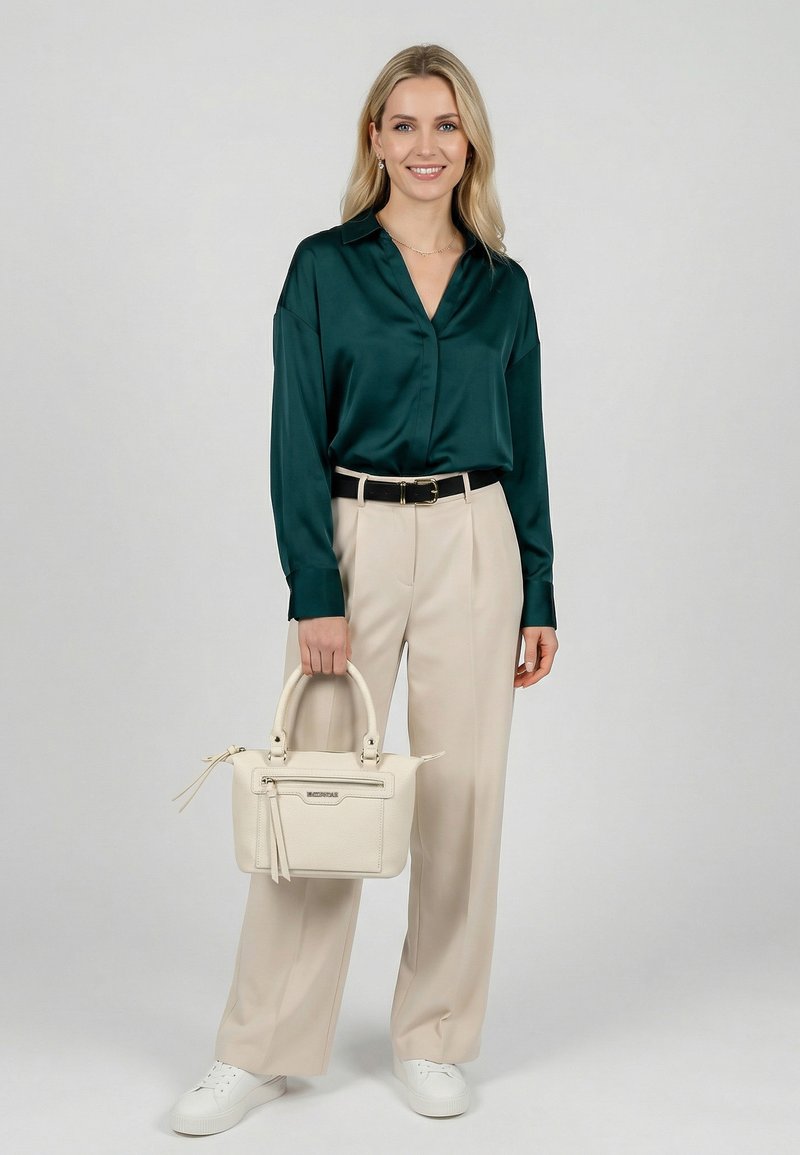 Hymyilevä nainen seisoo yllään tummanvihreä satiinipusero, beiget leveälahkeiset housut ja valkoiset lenkkarit, pitäen valkoista käsilaukkua yksinkertaista taustaa vasten.