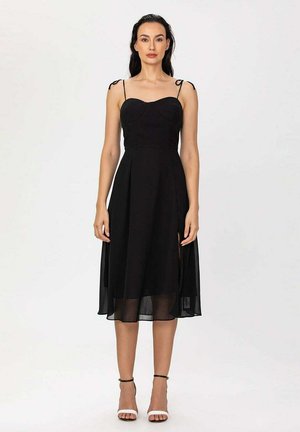 Femme debout portant une robe midi noire sans manches avec bretelles spaghetti et des sandales blanches à talons hauts sur un fond uni.