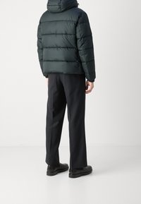 Persona che indossa un piumino nero, pantaloni dritti scuri e scarpe eleganti nere, in piedi di spalle su uno sfondo chiaro e semplice.