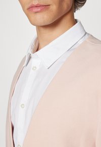 Hellrosa Strickjacke mit einem weißen Hemd darunter. Die Strickjacke hat ein V-Ausschnitt-Design und eine strukturierte Stoffoberfläche.