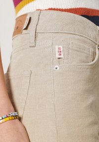 Pantalones de pana beige con una sutil textura acanalada, detalles de botón metálico y una pequeña etiqueta de la marca en la cinturilla.