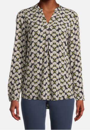 Vrouw draagt een blouse met lange mouwen met een grijze achtergrond en een wit, groen en beige geometrisch bloemmotief, gecombineerd met blauwe broek.