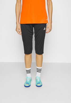 Personne portant un short de sport noir Adidas jusqu’aux genoux, des chaussettes blanches avec des rayures noires, des chaussures de sport turquoise et un t-shirt orange.