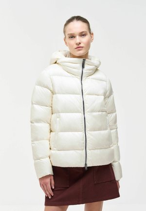 Frau trägt eine weiße Steppjacke mit Kapuze und einen dunkelbraunen Rock und steht vor einem einfarbigen weißen Hintergrund.