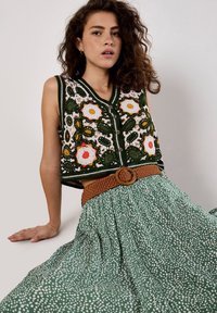 Crochet vest with floral patterns in green, yellow, and red, paired with a green polka dot skirt and a braided tan belt with a circular buckle.
