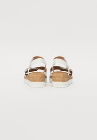 White sandals with brown cork-like wedge soles, adjustable straps, and textured detailing on the sole. Flat heel design.
