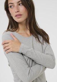 Maglietta grigia a maniche lunghe in un tessuto morbido e leggero, con scollo rotondo e maniche aderenti, abbinata a uno sfondo neutro.