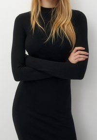 Blondynka z długimi włosami ubrana w dopasowaną czarną sukienkę z długim rękawem, z rękami skrzyżowanymi na piersiach na jednolitym białym tle.