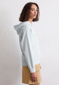 Lichtblauwe hoodie met een relaxte pasvorm, voorzien van een trekkoordkap en geribde manchetten. Draagt een tan rok, tegen een witte muur als achtergrond.