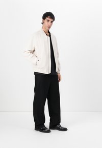 Light beige bomber jacket with elastic cuffs and hem, worn over a black top. Pair of black wide-leg pants and polished black shoes.