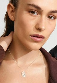 Collana con pendente d'argento, con un charm rettangolare, finemente progettato, sospeso a una catena sottile, indossato su pelle nuda con un indumento marrone.