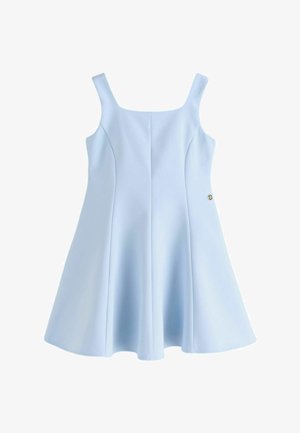 Junge Person, die ein ärmelloses, hellblaues ausgestelltes Kleid und weiße Sneakers mit beigen Sohlen auf einem grauen Boden trägt.