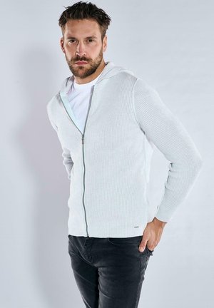 Hellgrauer, gerippter Zip-Hoodie mit weißem Innenfutter, langärmlig und in lässiger Passform. Getragen über einem weißen T-Shirt.