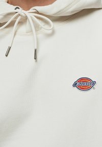 Sudadera blanca con cordón y puntas metálicas plateadas. Presenta un logotipo circular bordado en rojo, azul y naranja en el pecho.