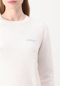 Maglione crema con scollatura rotonda, maniche lunghe e un sottile ricamo "2NDAY" in grigio chiaro sul petto. Tessuto morbido e strutturato.
