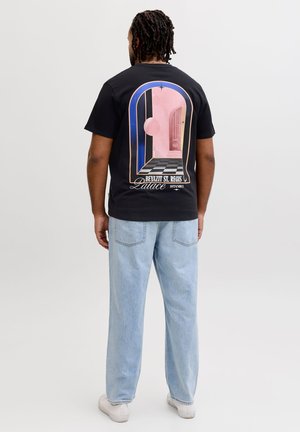 Homme aux cheveux tressés portant un T-shirt noir avec un arc géométrique coloré et du texte, un jean bleu clair et des baskets blanches, vu de dos.