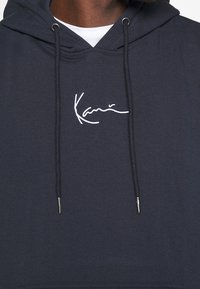 Navy blue hoodie made of cotton blend fabric, featuring a front kangaroo pocket and a white embroidered signature on the chest.