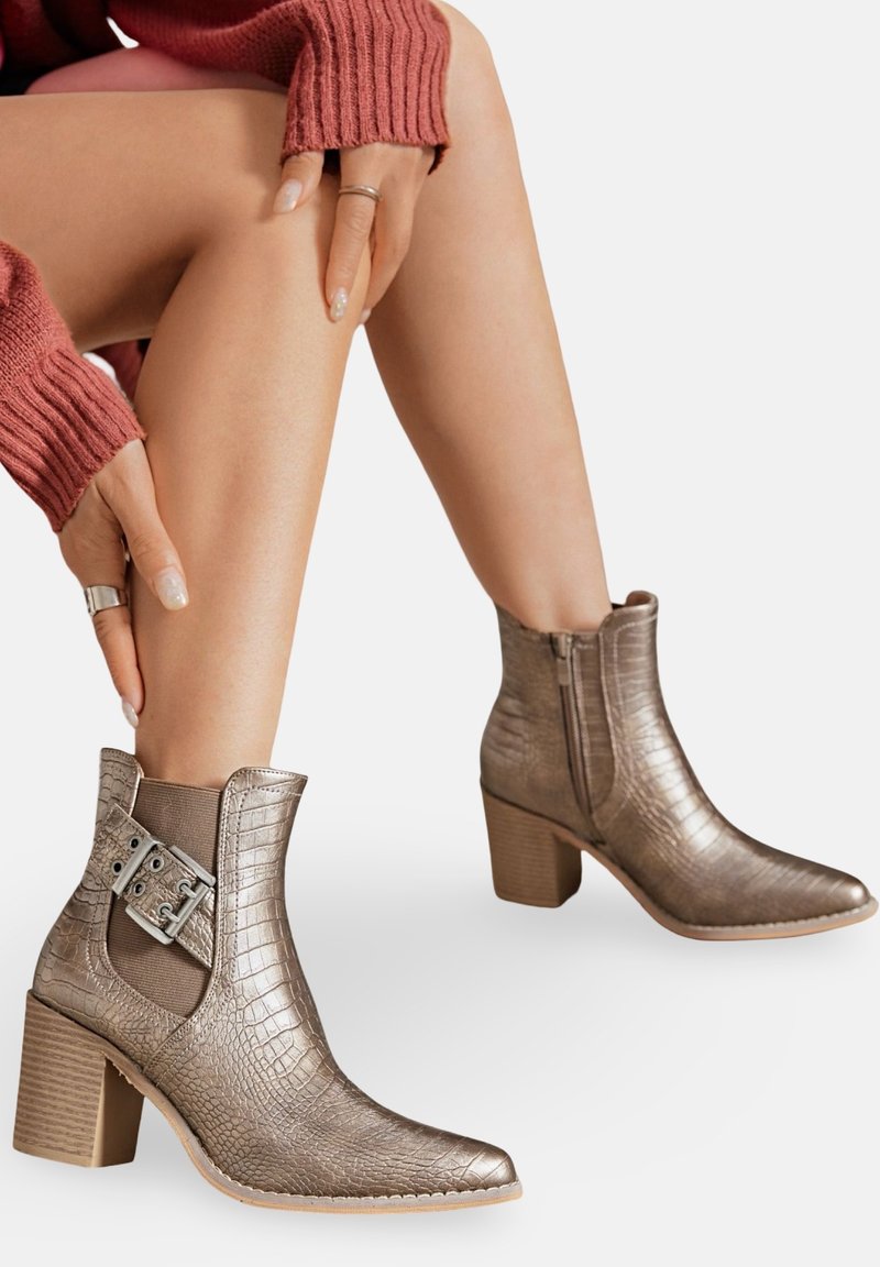 Femme portant des bottines métalliques à texture avec des talons bloc et des boucles, les mains reposant sur des jambes nues, devant un fond uni.