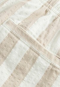 Tissu rayé beige et blanc avec une surface texturée, présentant une couture visible et un détail de poche à rabat. Le matériau semble doux et léger.