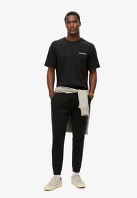 Zwarte t-shirt met korte mouwen en wit logo, gecombineerd met zwarte joggers en een grijze trui die om de taille is geknoopt. Grijze en beige sneakers maken de look compleet.