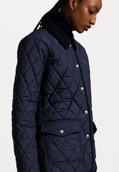 Navyblaue gesteppte Jacke mit einem Cordkragen, Druckknöpfen und einer Fronttasche. Zu den Details gehört ein kleines rotes Logo auf der Brust.