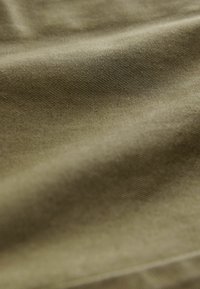 Tissu vert olive avec une texture côtelée subtile, montrant des plis lisses et un doux éclat sous la lumière, idéal pour le rembourrage ou les vêtements.