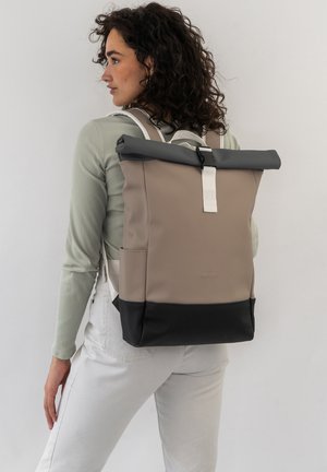 Rolled-top backpack in beige, gray, and black. Features a sturdy strap closure, textured surfaces, and side pocket. Includes white accents.