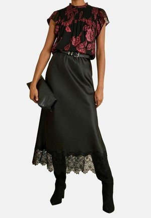 Vrouw draagt een zwarte satijnen midi-rok met kanten zoom, een rood-zwarte bloemenblouse, een zwarte riem, laarzen en houdt een zwarte clutch vast.