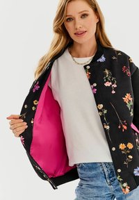 Czarna bomberka w kwiaty z kontrastowym różowym wnętrzem, zdobiona różnymi wzorami kwiatowymi i zapinana na zatrzaski.