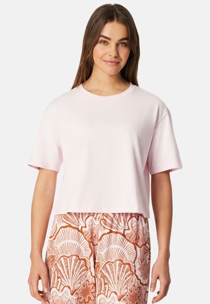 Jonge vrouw die een effen lichtroze T-shirt met korte mouwen draagt en een broek met hoge taille met grote oranje zeeschelpenpatronen.