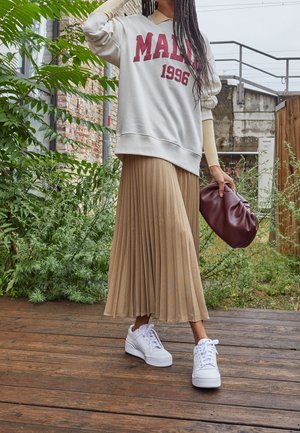 Weißer Sweater mit burgunderrotem Text, beigefarbener Faltenrock, weiße Sneaker und eine burgunderrote Clutch-Tasche, vor grüner Umgebung und Holzterrasse.