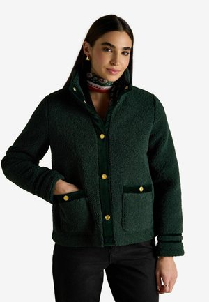 Grüne Fleecejacke mit strukturierter Oberfläche, Samt-Akzenten, goldenen Knöpfen und Vordertaschen. Kombiniert mit einem gemusterten Schal darunter.