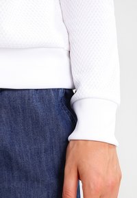 Sweatshirt blanc texturé avec un motif en diamond et des poignets côtelés, associé à un jean foncé. Accent sur le matériau et le design.