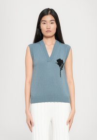 Ermeløs teal genser med V-halsdesign, ribbestrikkede kanter og en svart blomsterbroderi detalj på fronten.