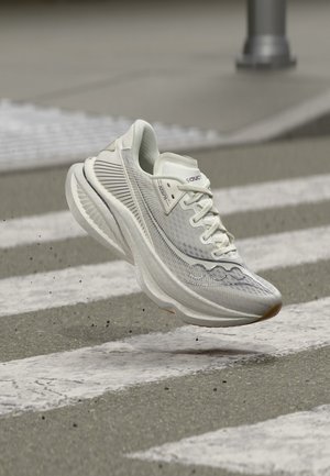 Άσπρο παπούτσι για τρέξιμο Saucony αιωρείται πάνω από διάβαση πεζών σε ασφάλτινο δρόμο με μικρά θραύσματα πεταμένα γύρω.