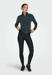 Dug madavne majice s dugim rukavima u tamnozelenoj boji s crnim leopard uzorkom, uparen s crnim tajicama koje imaju suptilan životinjski uzorak i crnim sportskim cipelama.