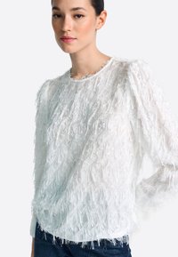 Sieviete tērpusies baltā, garu piedurkņu teksturētā augšdaļā ar spalvu rakstu, kombinējot to ar tumši zilām džinsu biksēm, atrodoties pret vienkāršu fonu.