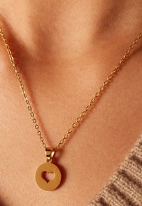 Gouden ketting met een ronde hanger met een hart uitsnede, hangend aan een fijne schakelkettinkje, rustend tegen een huidskleurige achtergrond.