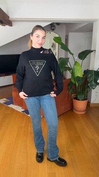 Mladá žena s copom, oblečená v čiernom svetri GUESS a modrých džínsoch, stojí v obývacej izbe s oranžovou pohovkou a veľkou kvetináčovou rastlinou za ňou.