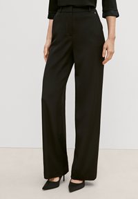 Czarne spodnie o szerokich nogawkach z gładką teksturą, z bocznymi kieszeniami i dopasowanym krojem. Noszone z czarnymi czółenkami w szpic.